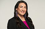 A woman with long dark hair wearing a suit jacket and pink shirt. She has long blue earings and is smiling.