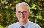 White man with short white hair and dark, wide-framed glasses staring ahead with a sile.