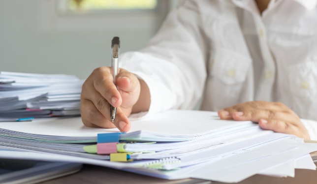 Teacher hand is holding pen for checking student homework assignments on desk in school. Unfinished paperwork stacked in archive with color paper and binder paper clips. Education and business concept