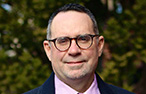 A white man (Mark Robertson) wearing glasses and looking at the camera with a slight smile. He wears a purple collared shirt and suit jacket.